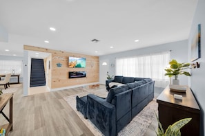 A modern living room with a sectional sofa, a flat-screen TV mounted on a wooden wall, a dining area with a table and chairs, and a staircase leading to another level. - Waterfront Gulf-access home w/heated pool, boat lift, kayaks, & stunning sunsets (Cape Coral)