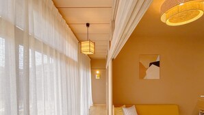 Une pièce avec un plancher en bois, un canapé jaune, une table en bois, des rideaux légers et un luminaire suspendu.