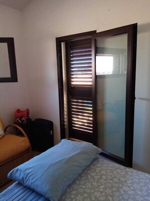 Una habitación con cama, una silla, un maletero y una puerta corredera con persianas.