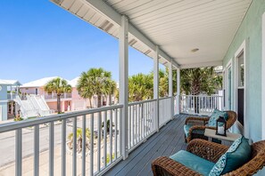A covered porch with wicker furniture and a railing, overlooking a street with buildings and palm trees. - 🔥 🏖️ Gone To The Beach: Firepit, Community Pool + Steps to Navarre Beach! (Navarre Beach)