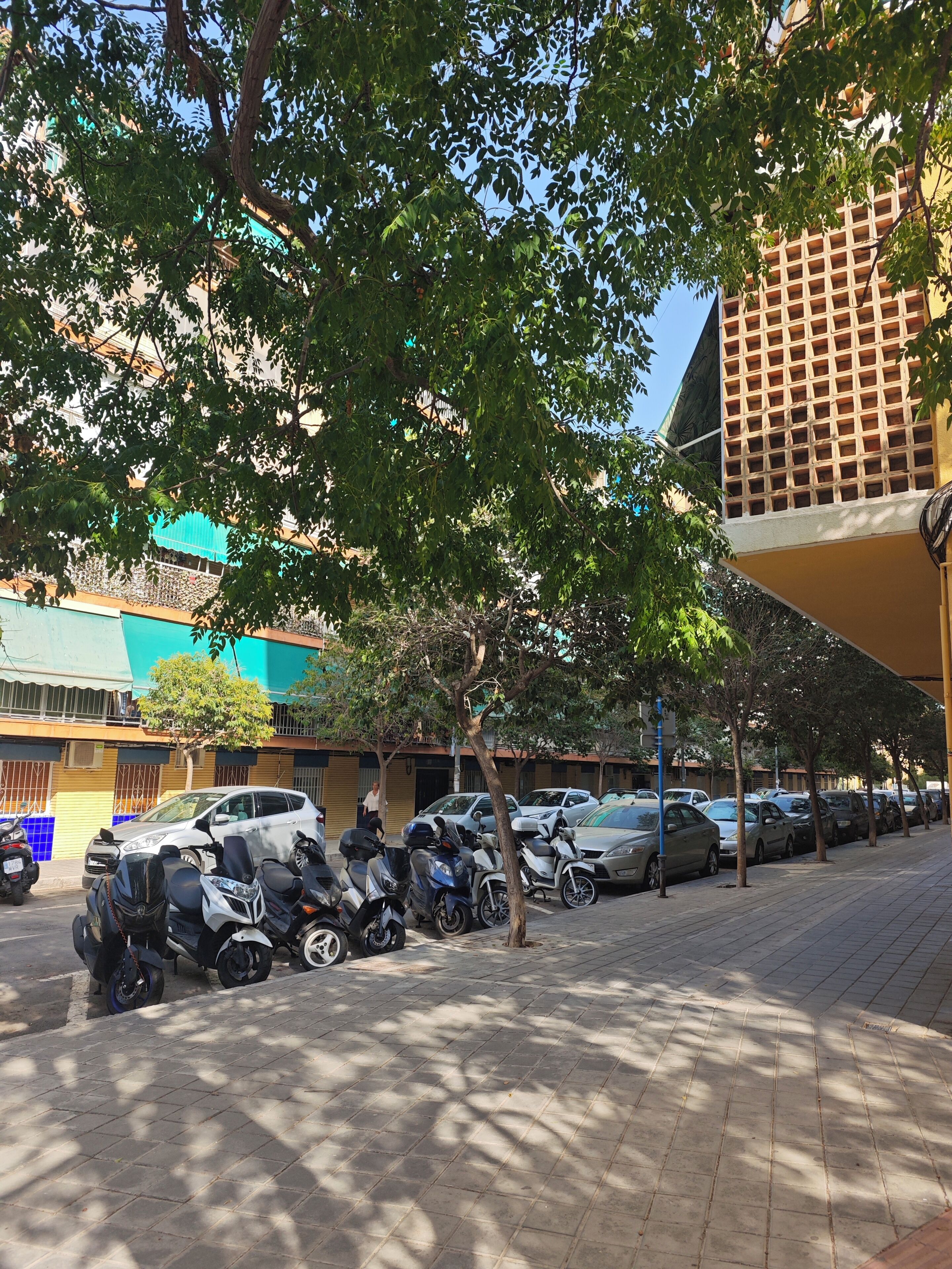 Un espace de stationnement où sont garés plusieurs scooters et voitures, un trottoir bordé d’arbres et un bâtiment doté d’une façade à motifs distinctifs.