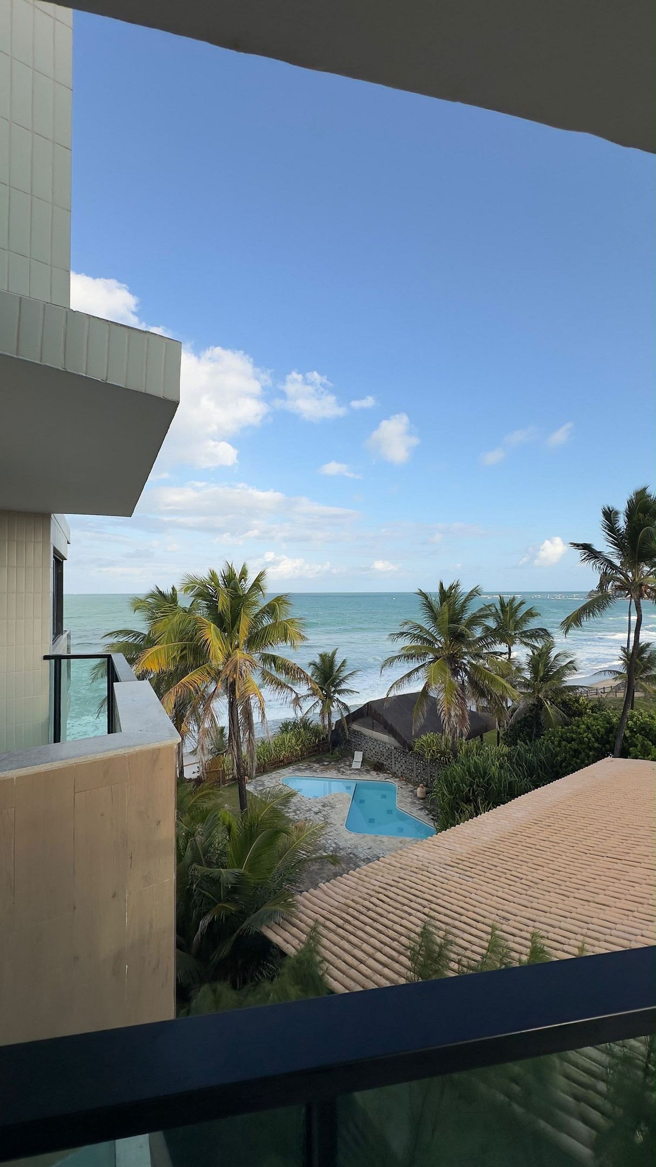 A pool area with a view of the ocean, surrounded by palm trees and a tiled roof.