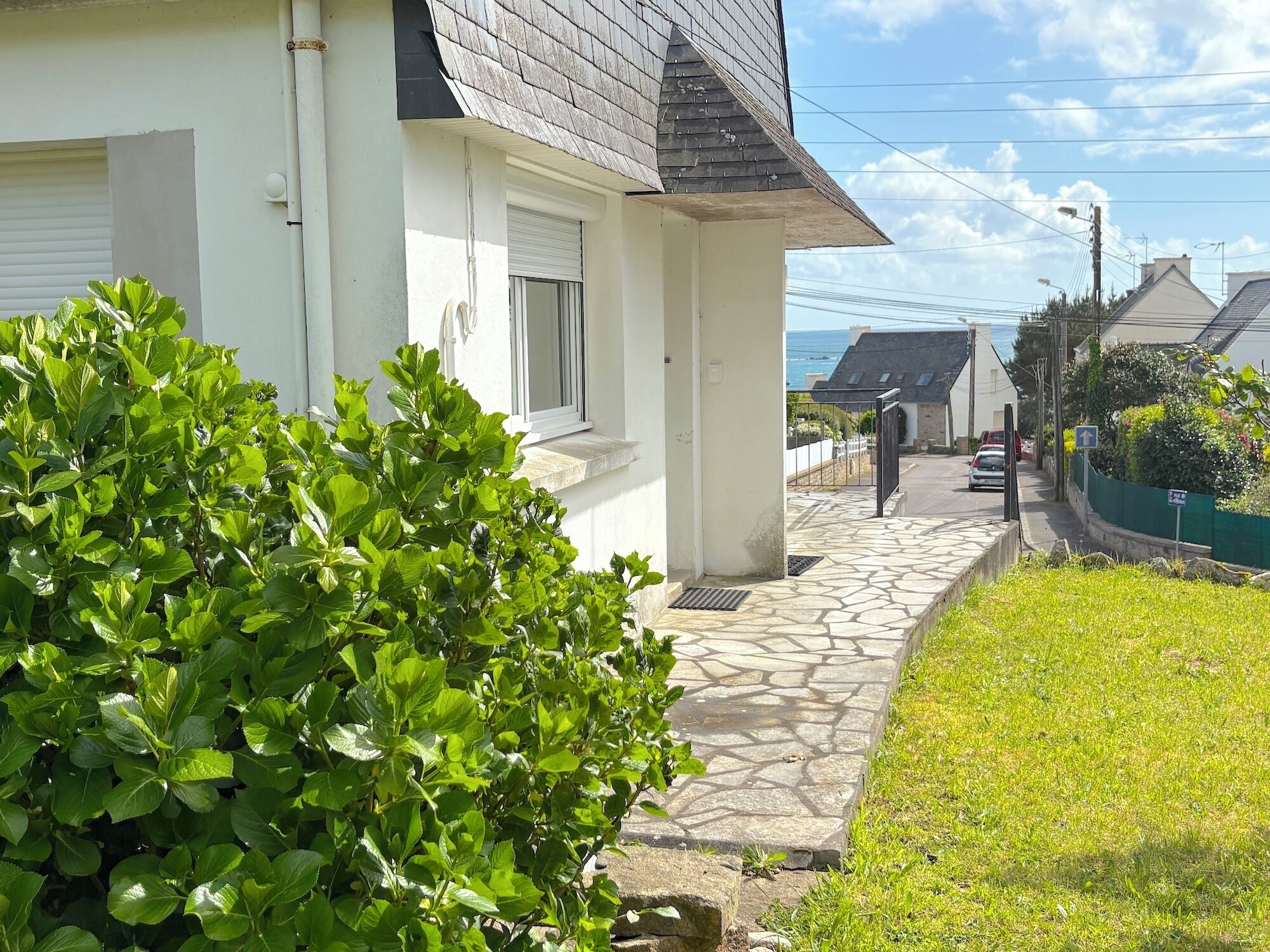 Une maison située sur la côte, avec un chemin de pierre menant à une entrée fermée par un portail, une façade blanche et une vue sur la mer.
