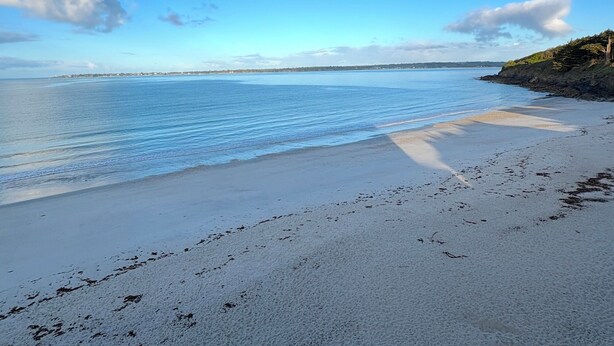 Ein Strand mit klarem, blauem Wasser, einem sandigen Ufer und einer fernen Küstenlinie mit Gebäuden.