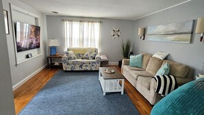 A living room with a TV, sofa, coffee table, and a large window with curtains. -  Charming 3 bed/2 bath house in Rock Hall with a 3-min walk to restaurants/town. (Rock Hall)