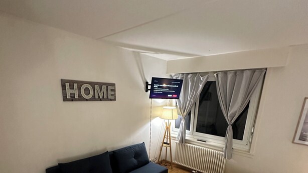 A small, cozy room with a dark-colored sofa, a bed with a patterned bedspread, a standing lamp, and a TV on the wall. - Central and comfortable apartment for two or three people (Lausanne)