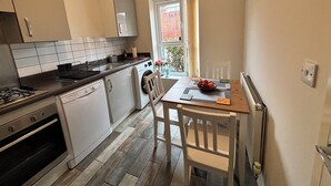 A compact kitchen with a stove, dishwasher, and a dining area with a table and chairs. - Cozy Home Walk to Anfield and Liverpool Attractions (Liverpool)