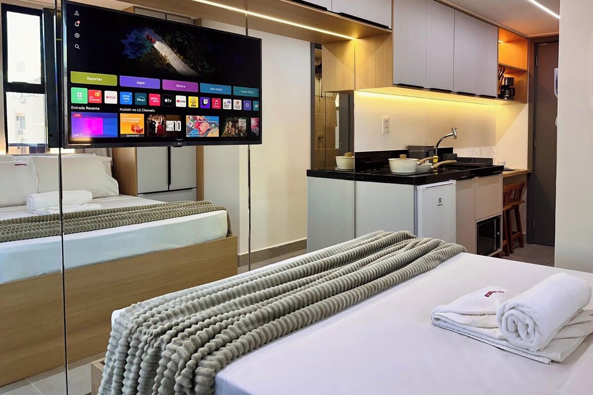 Moderni hotellihuone, jossa on sänky, televisiossa näkyy kanavavalikko, huoneessa on pieni keittiö sekä kylpyhuone.