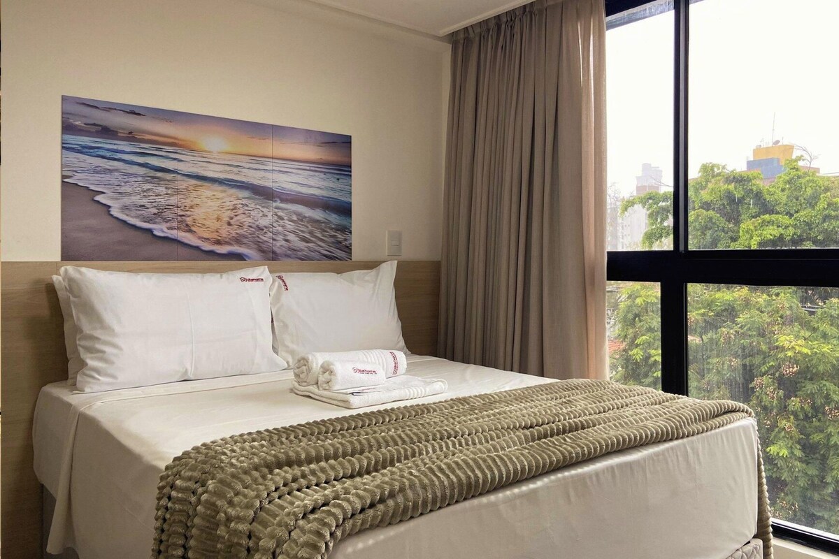Una habitación de hotel con cama, un cuadro de una escena de playa y vistas a un paisaje urbano a través de una ventana.
