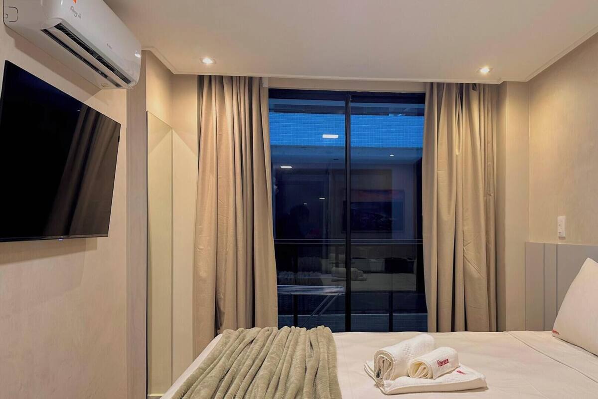 Una habitación de hotel con cama, televisor, ventana con cortinas y aire acondicionado.