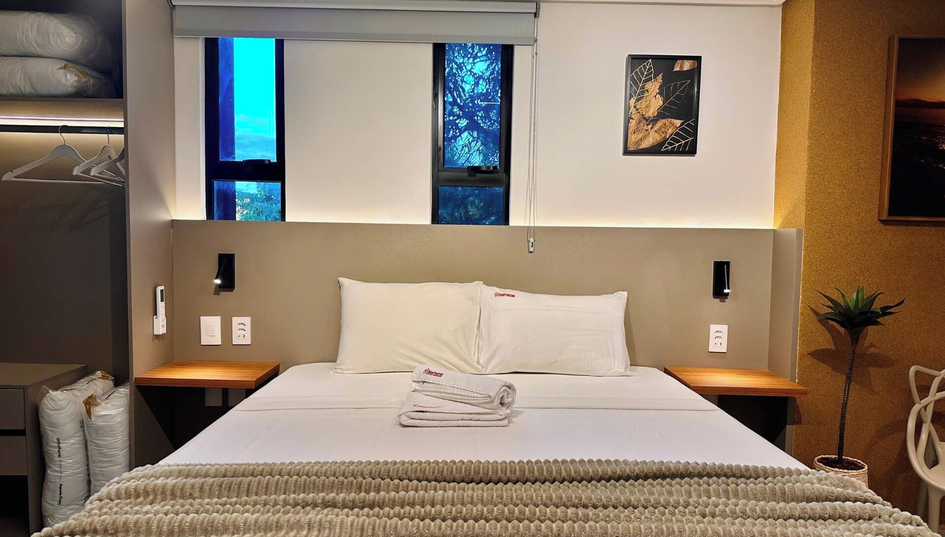Una habitación de hotel con cama, dos mesitas de noche, ventana con vistas y una obra de arte colgada en la pared.