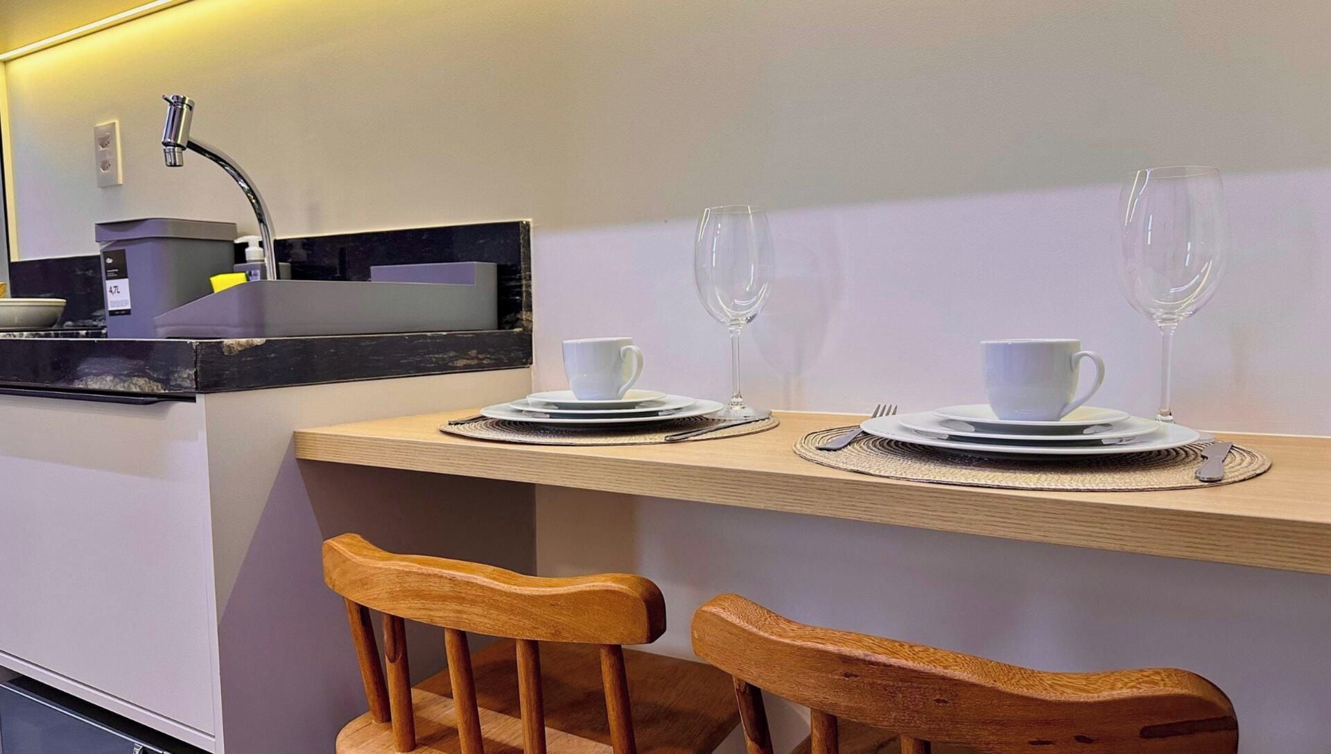Uma pequena cozinha compacta com pia, micro-ondas e cafeteira. Há duas cadeiras de madeira e uma mesa posta com pratos, xícaras e uma taça de vinho.