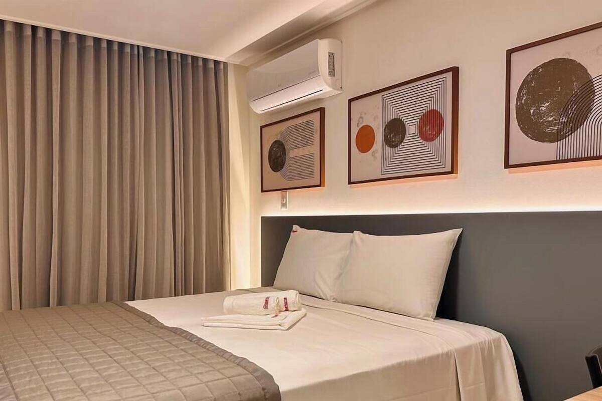 Una habitación de hotel con cama, cortinas, aire acondicionado y cuadros en la pared.
