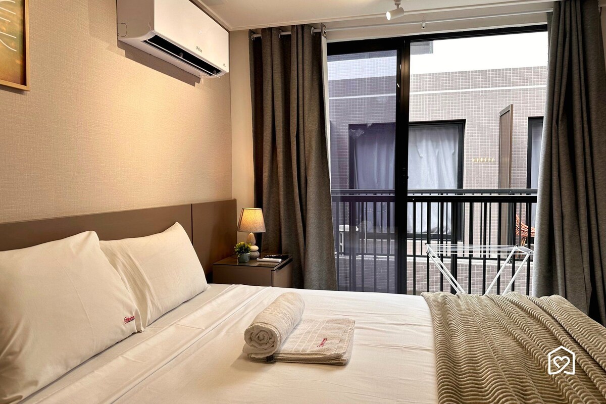 Una habitación de hotel con una cama, una mesita de noche con lámpara, una ventana con cortinas y vistas al exterior.