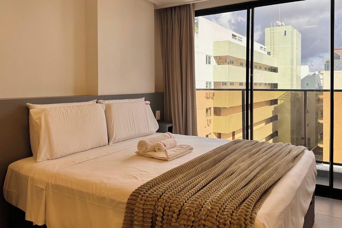 Habitación de hotel con una cama grande, vistas de la ciudad a través de ventanas de piso a techo y un balcón con vistas a los edificios.