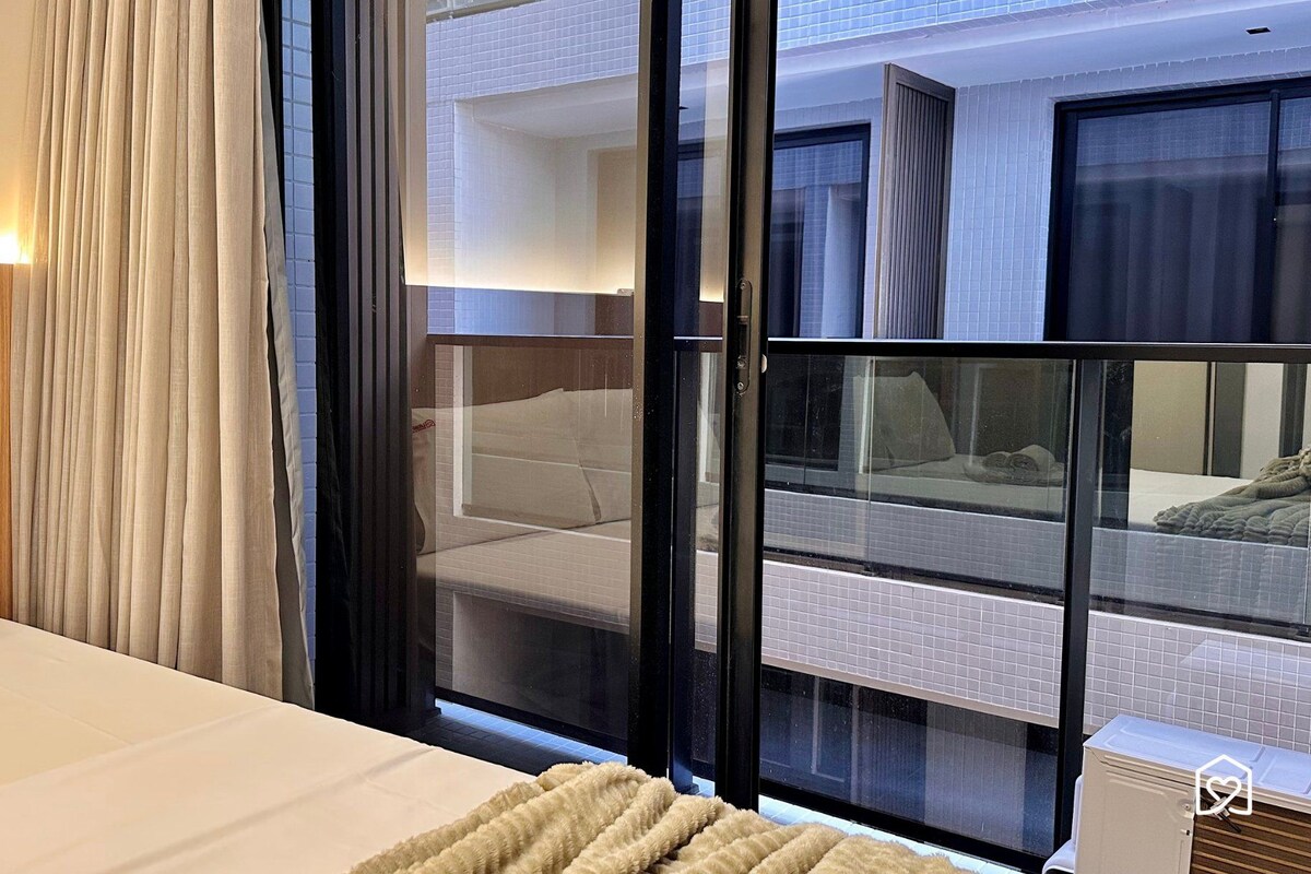 Una habitación de hotel con una cama, una ventana que da a otra habitación y una cortina.