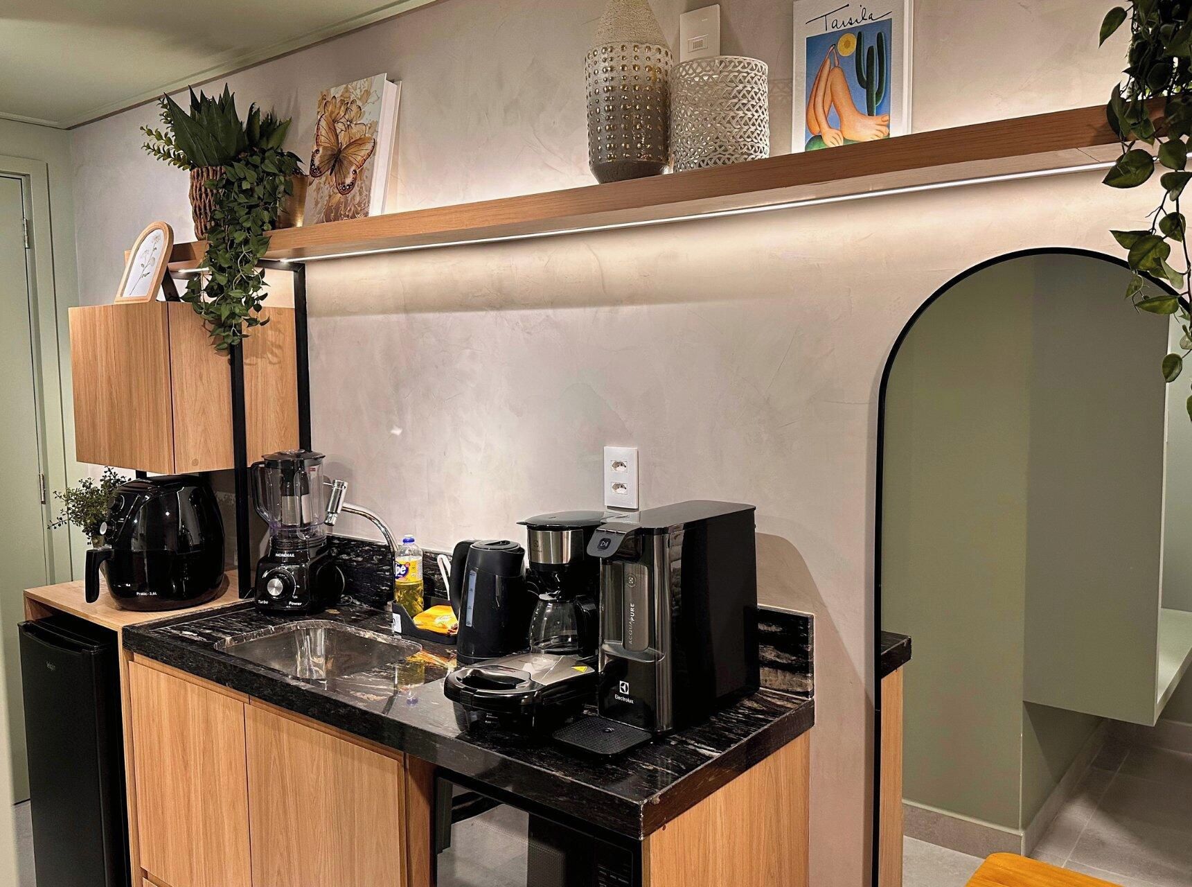 Cozinha moderna com bancada preta, cafeteira, liquidificador e uma geladeira compacta.