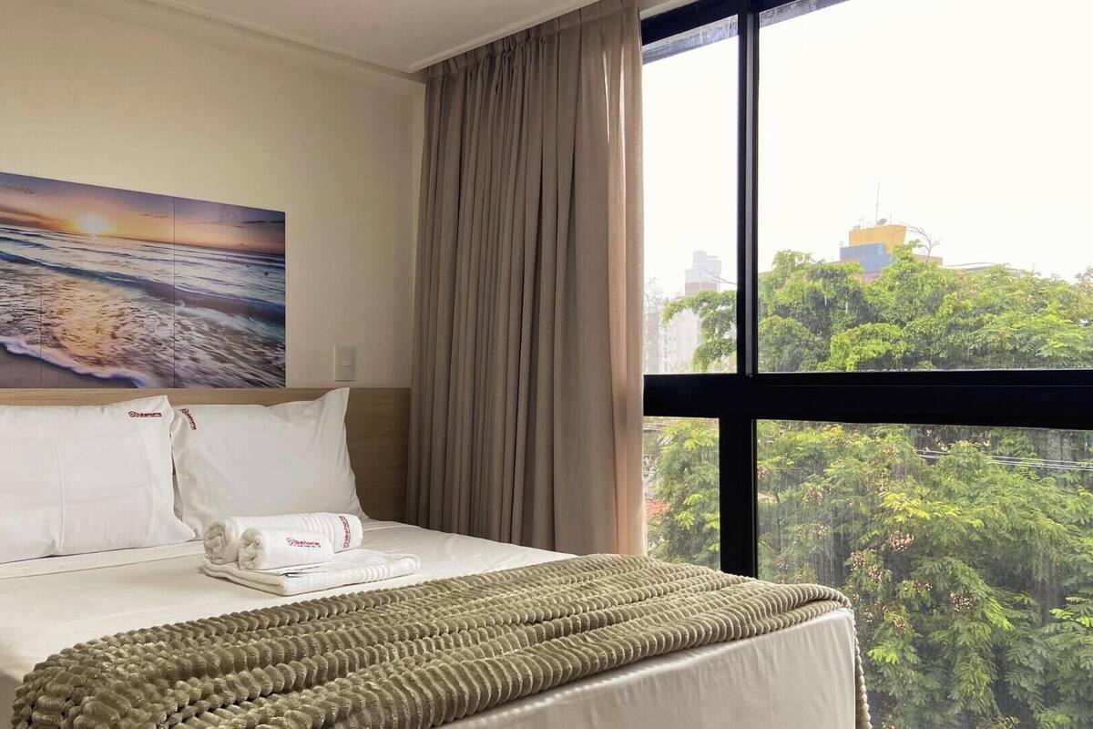 Una habitación de hotel con una cama, un cuadro de una playa, una ventana con vista a árboles y edificios, y una cortina.