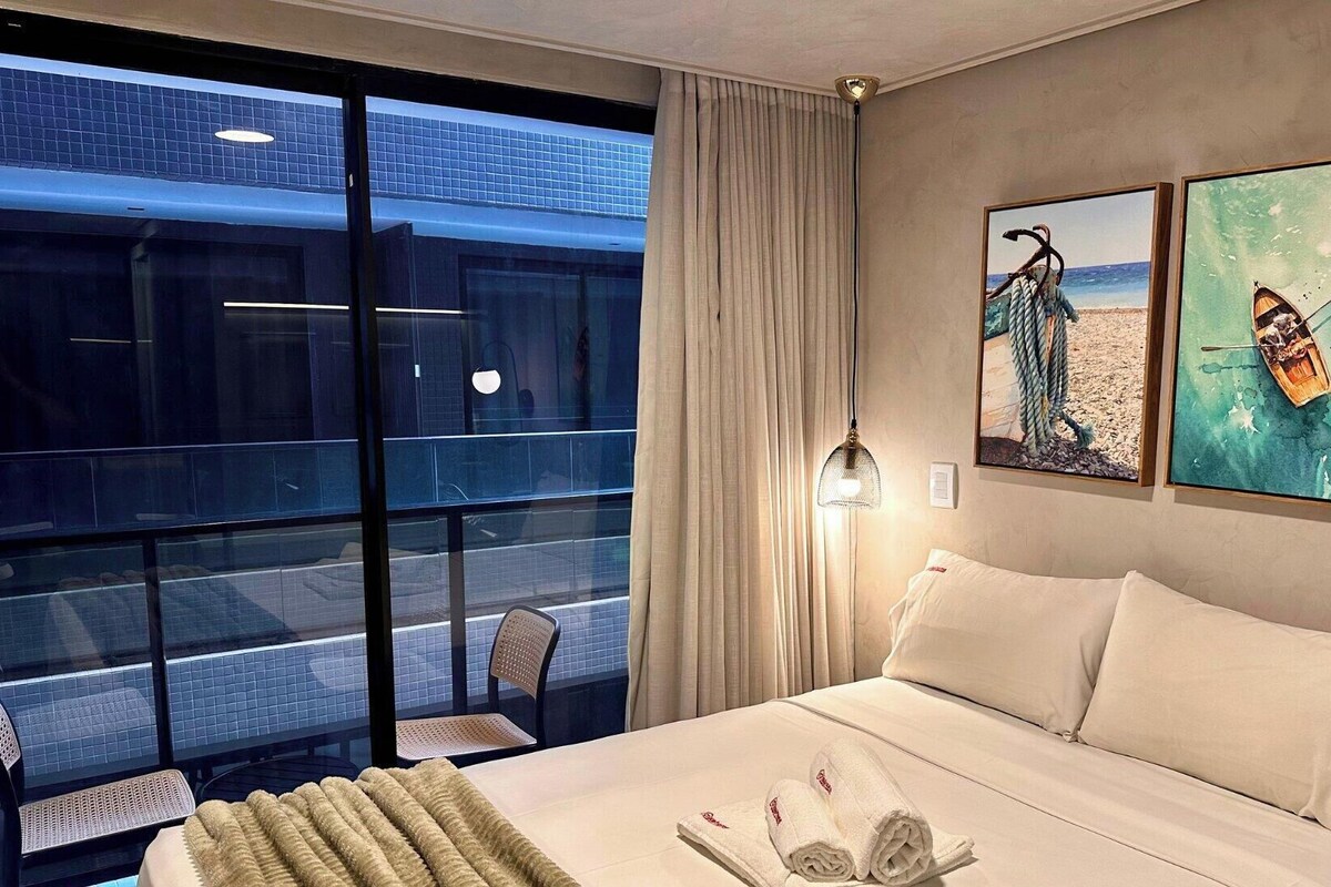 A hotel room with a bed, a chair, a table, and a view of a building through a window.