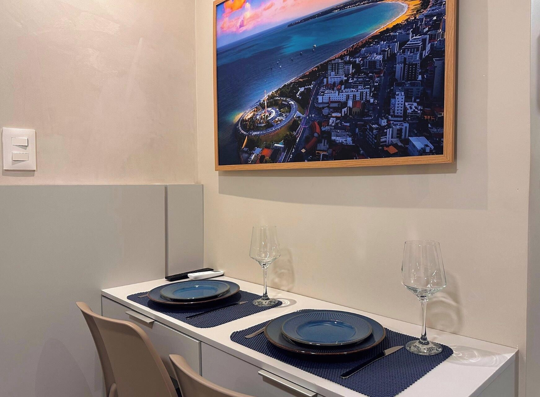 Uma sala de jantar com mesa posta para dois, uma pintura de um cenário costeiro na parede e vista para o mar.