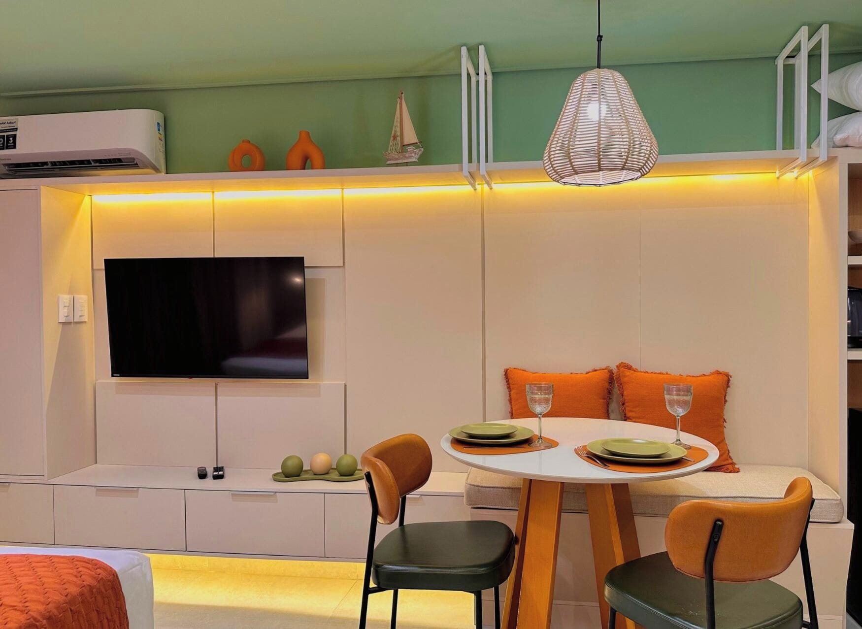 Quarto compacto com cama, mesa de jantar com cadeiras, TV de tela plana na parede e luminária no teto.