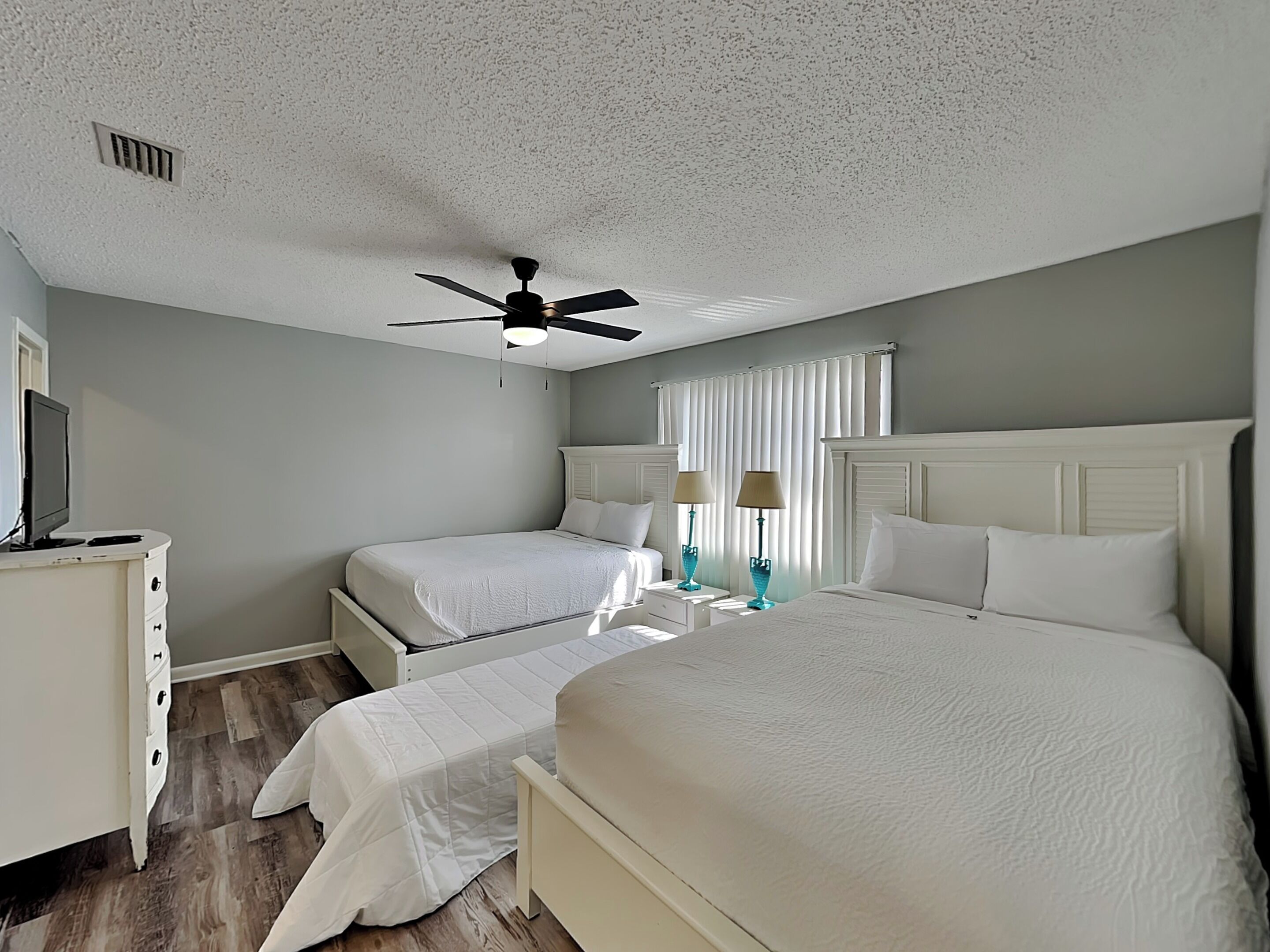 Une chambre d’hôtel comprenant un lit, deux tables de chevet, un ventilateur de plafond, une fenêtre avec des rideaux et un téléviseur posé sur un meuble.