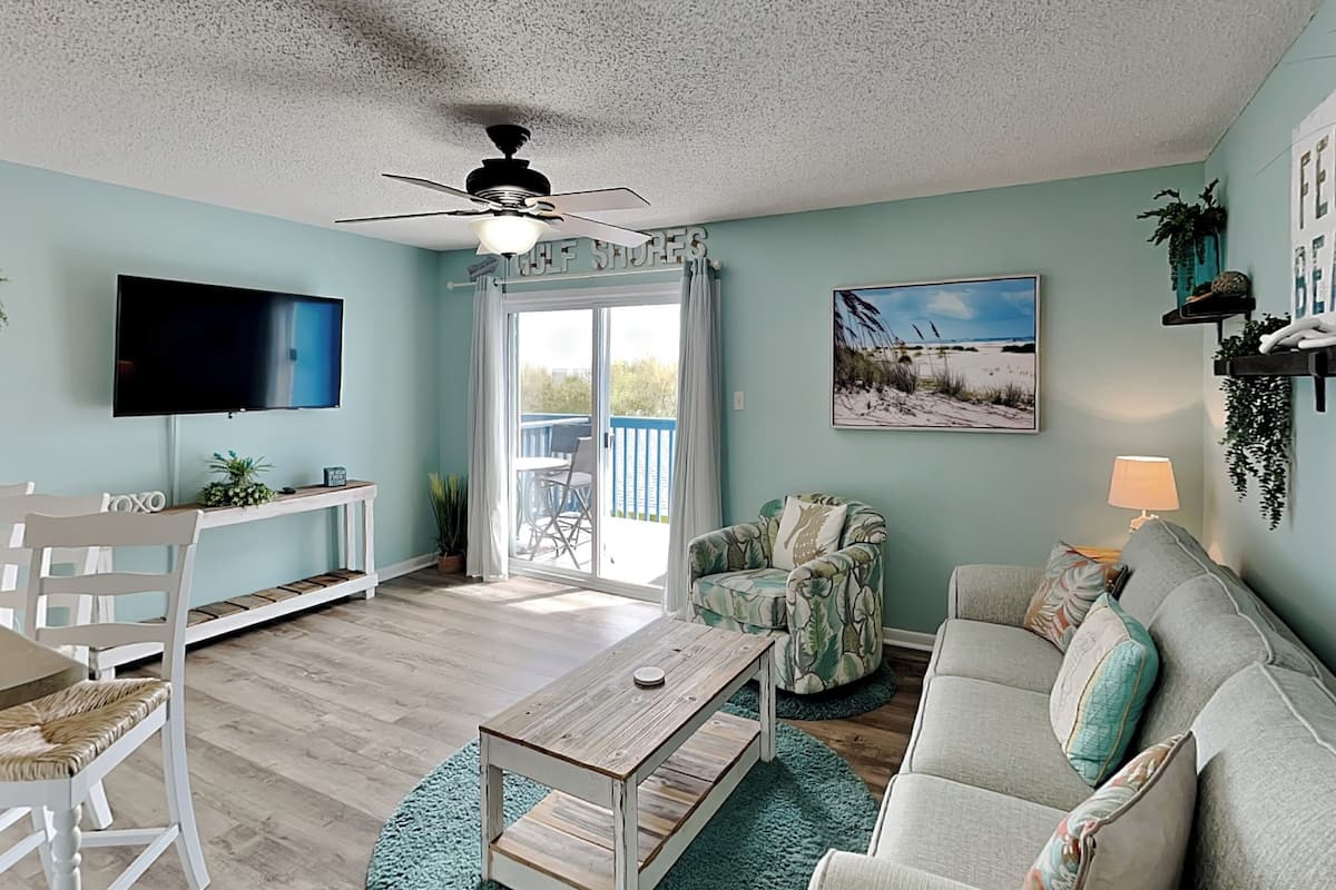 A living room with a TV, a ceiling fan, a sofa, a chair, a coffee table, and a balcony with a view.