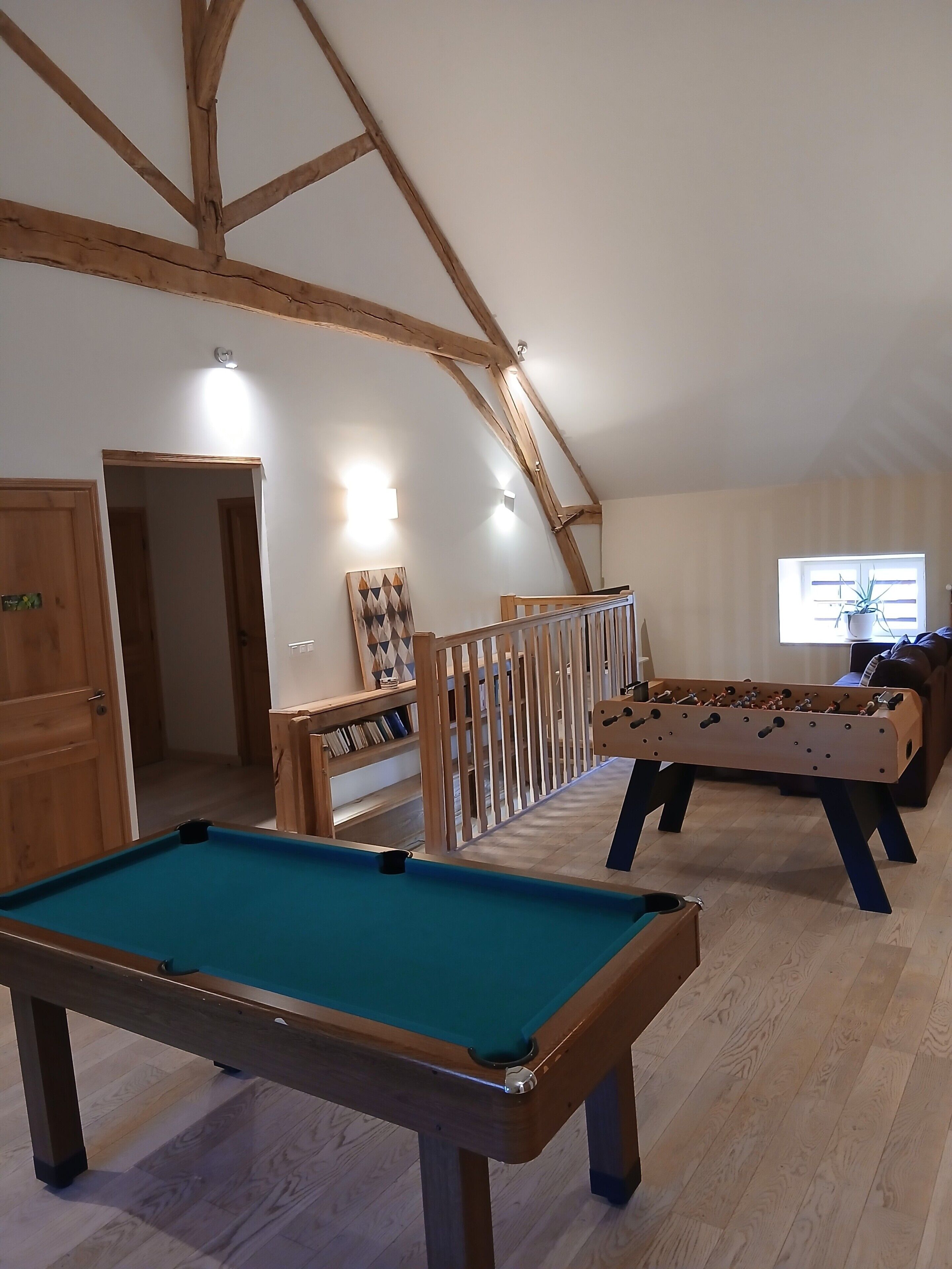 A room with a pool table, a foosball table, wooden beams on the ceiling, and a bookshelf.
