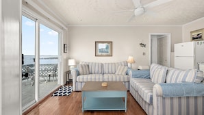 A living room with a sofa, a coffee table, a ceiling fan, a refrigerator, and a view of the ocean. - Bayfront Condo with Great Location in Ocean City (Ocean City)