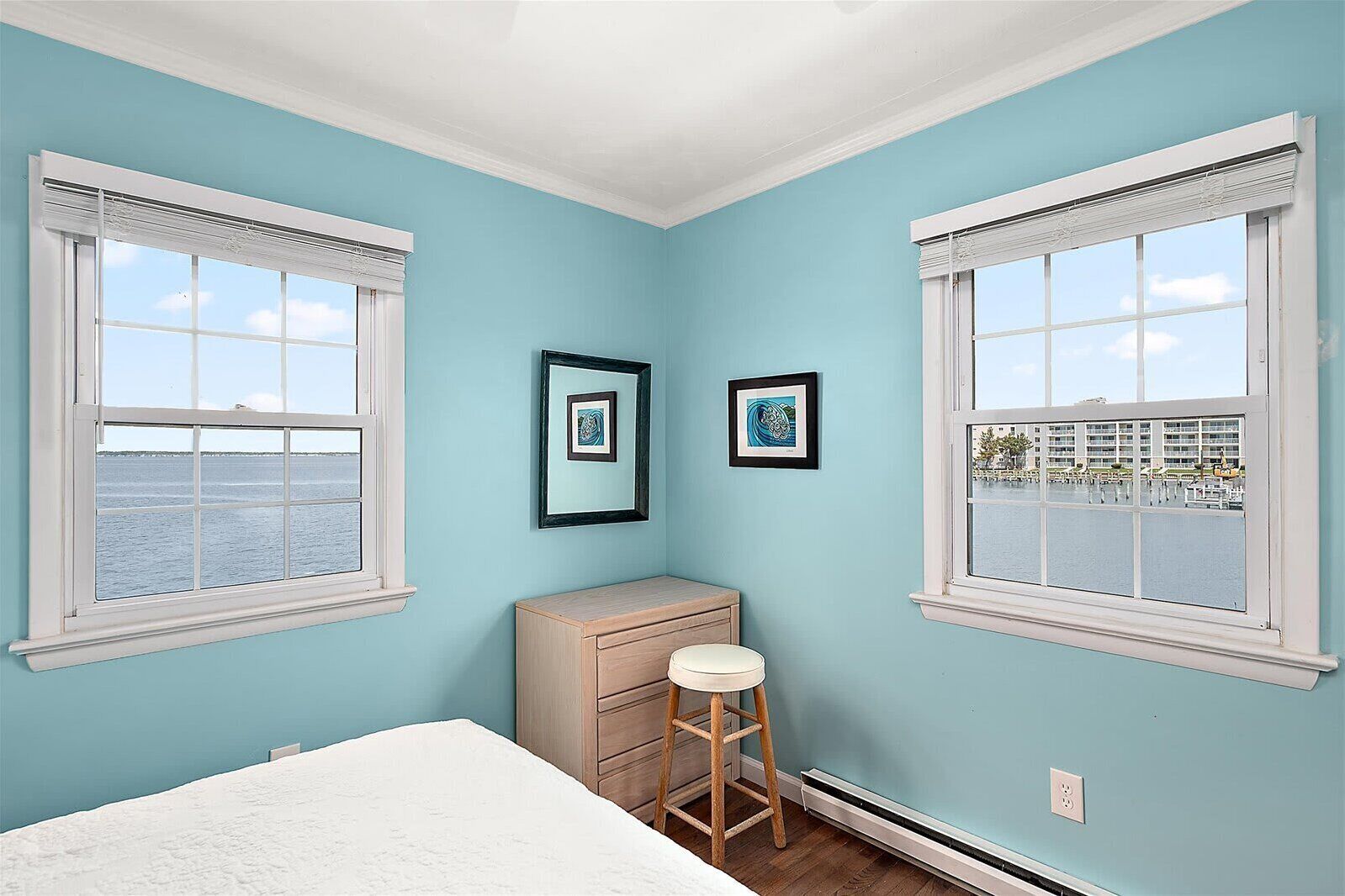 Una habitación con dos ventanas, una cama, un tocador, un taburete y dos cuadros en la pared.