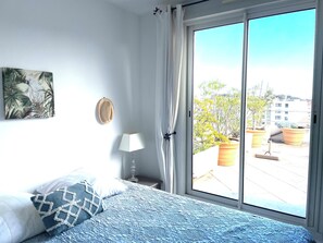A bedroom with a bed, a wall-mounted lamp, a hat, a painting, and a view of the outside. - Apartment with a large terrace (Cavalaire-sur-Mer)