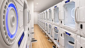 A row of white, modern, enclosed pods with digital displays, arranged in a linear fashion along a wooden floor. - Beddy Hostels (Madrid)