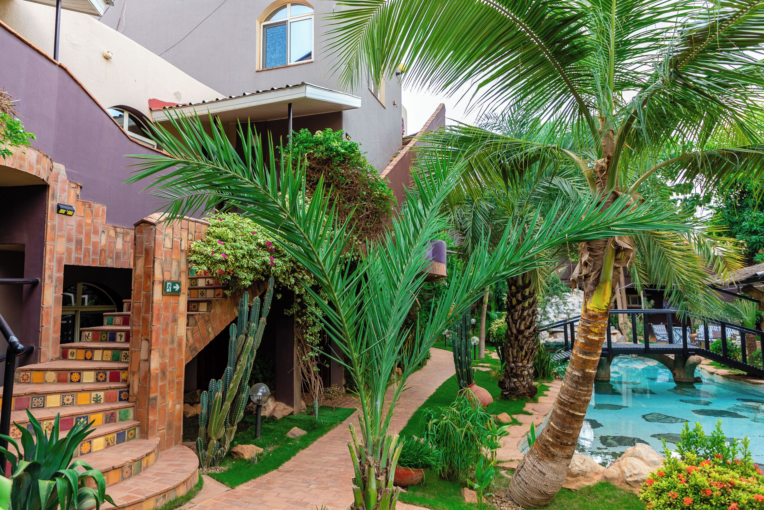 A courtyard with a pool, palm trees, and a staircase leading to a building with a colorful tile design.