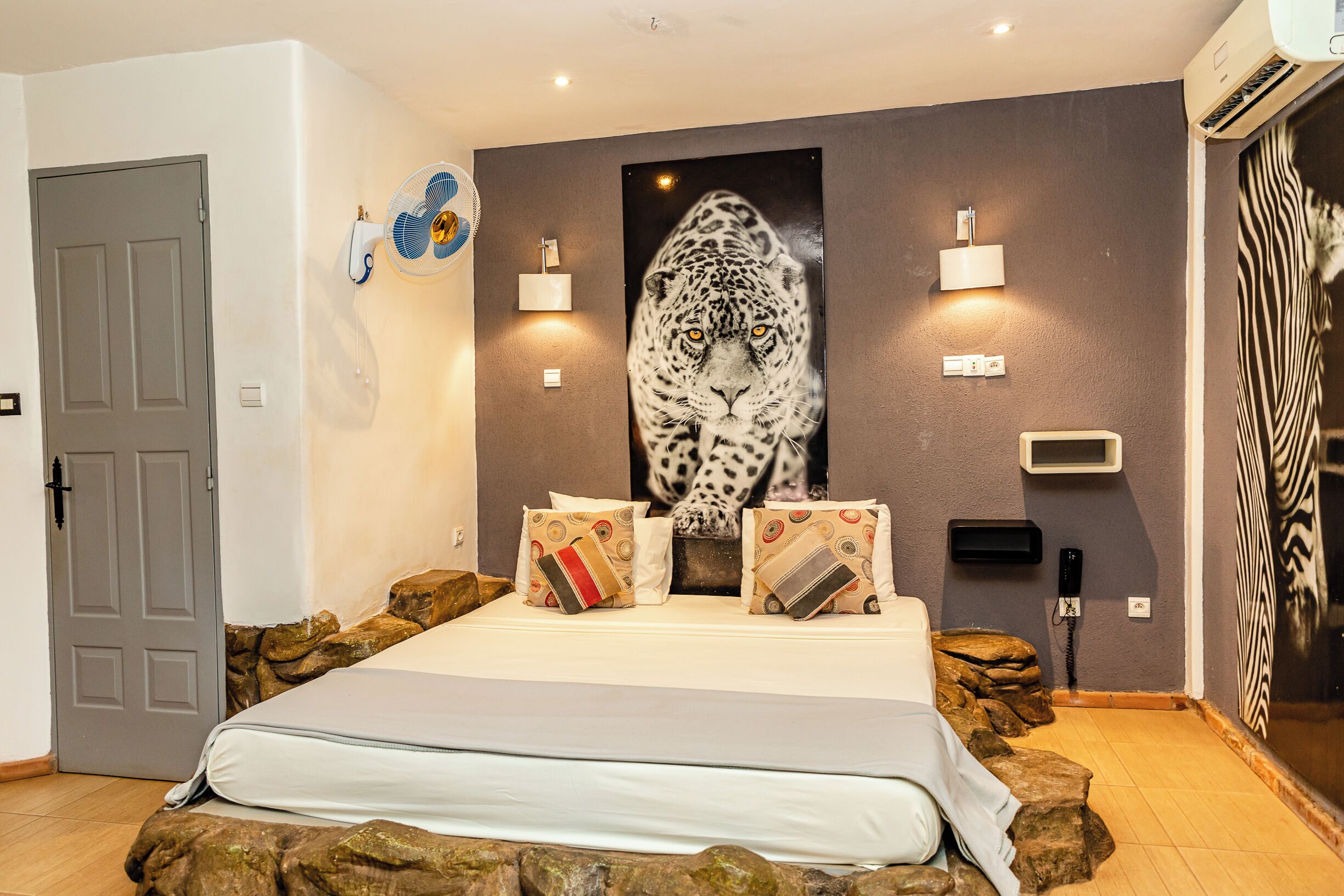 Ein Schlafzimmer mit einem Bett, Kissen, einem Wandventilator, einer Wandlampe und einem großen Tigerbild.