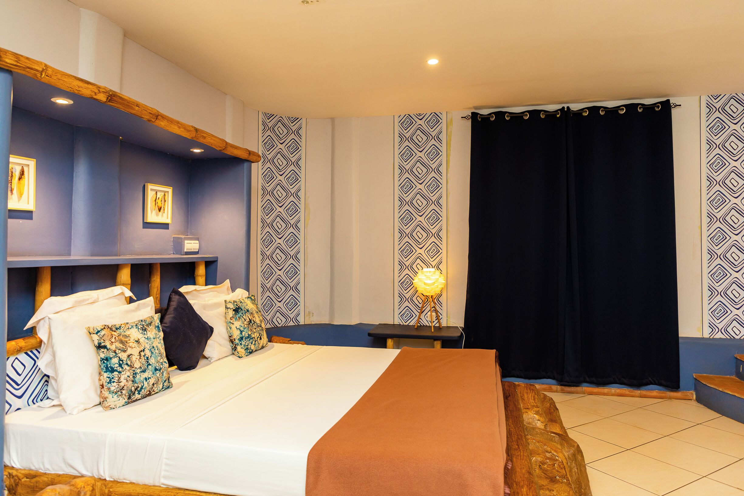 A bedroom with a large bed, a bedside table with a lamp, patterned curtains, and a wooden headboard.