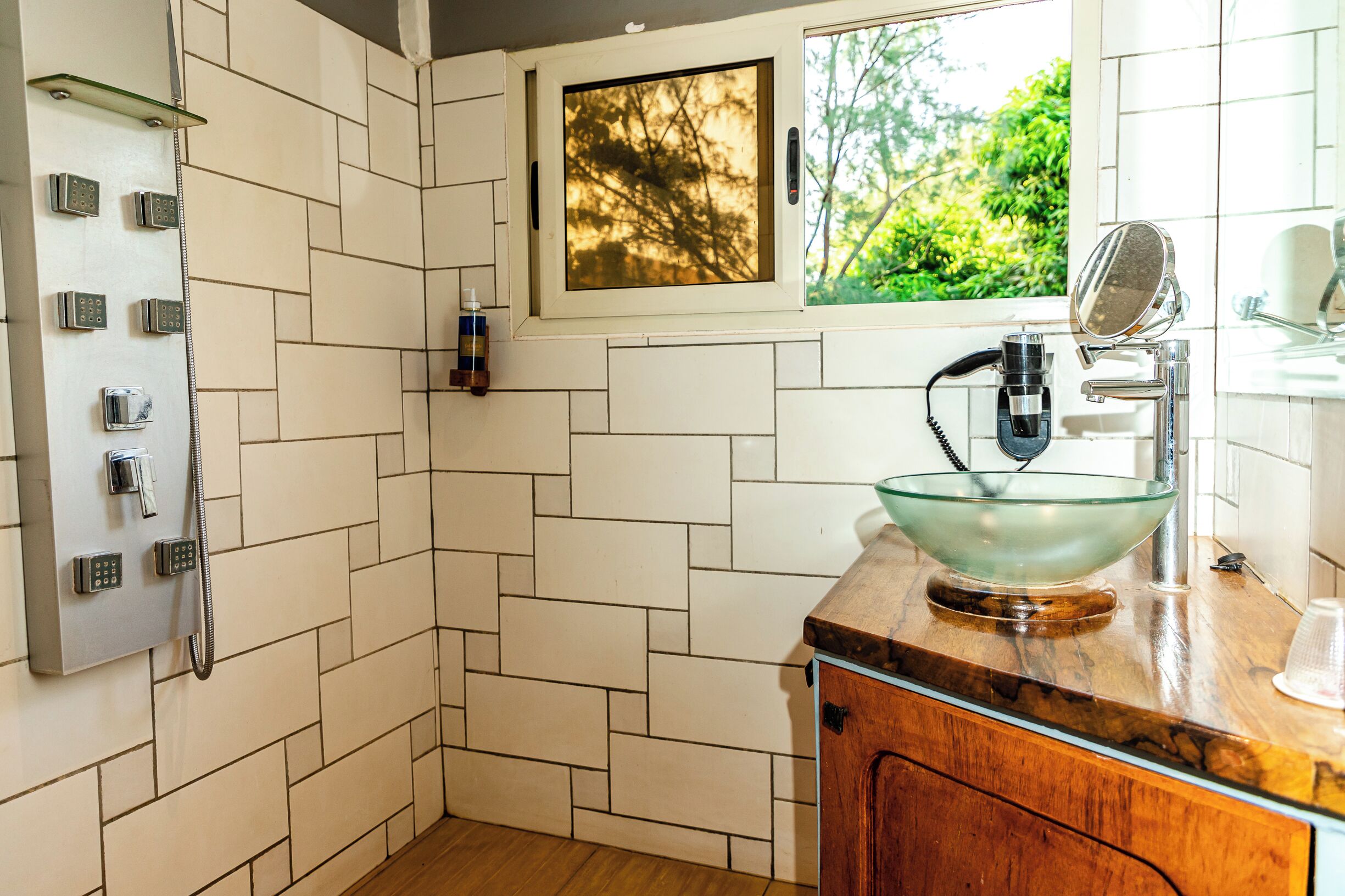 A bathroom with a shower, a glass bowl sink, a mirror, and a window with a view of greenery.