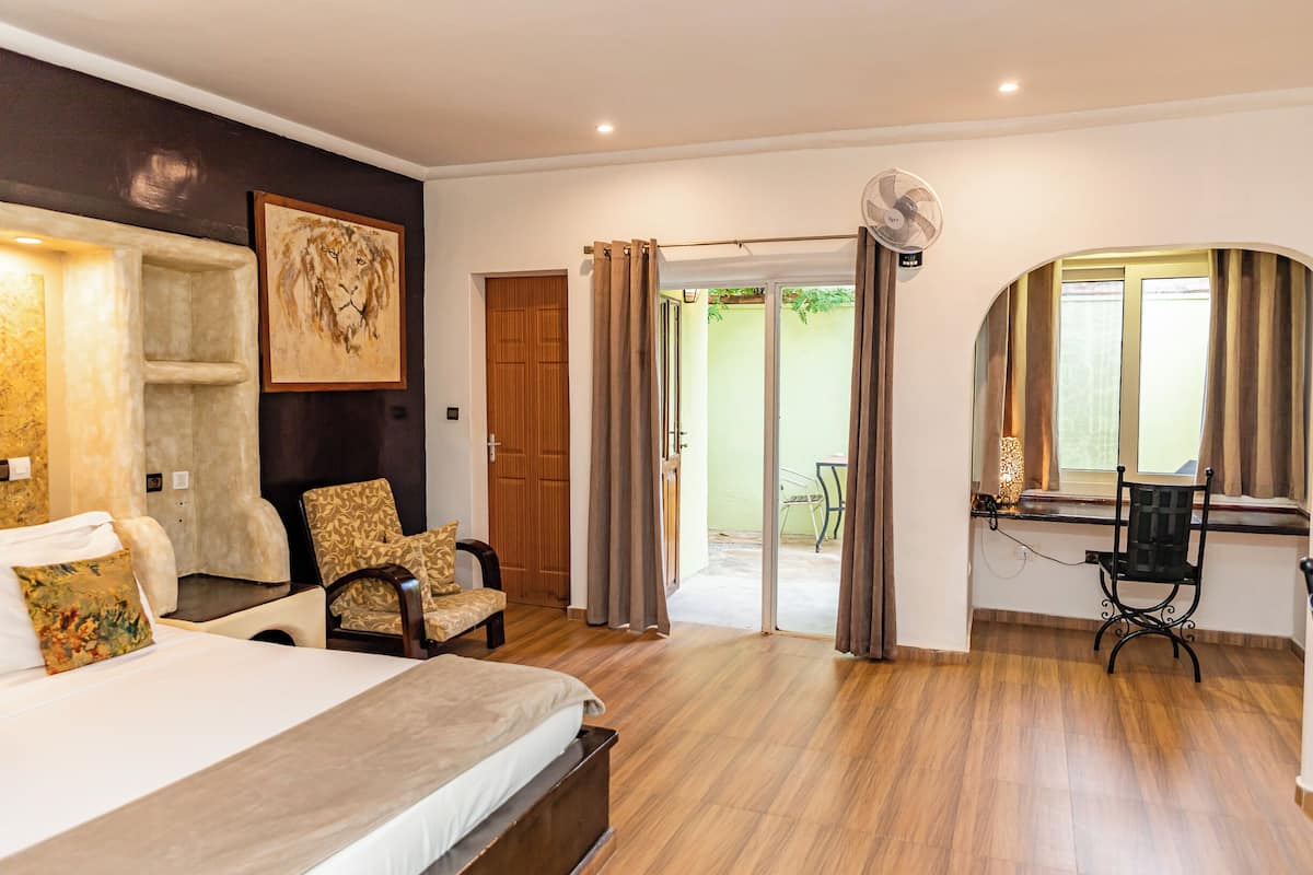 A bedroom with a bed, two chairs, a desk with a chair, a fan, and a sliding glass door leading to a balcony.