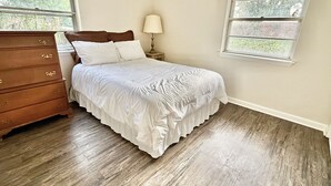 Un dormitorio con un aparador de madera, una cama con ropa de cama blanca, una mesita de noche con una lámpara y dos ventanas.
