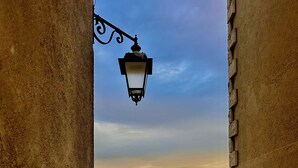 Une ruelle étroite bordée d’un mur de briques, avec une lanterne en métal, et d’où l’on aperçoit des champs lointains sous un ciel dégagé.