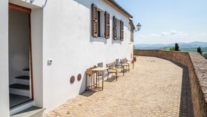 Un bâtiment blanc avec des volets en bois, une terrasse en briques et une vue sur des montagnes lointaines.