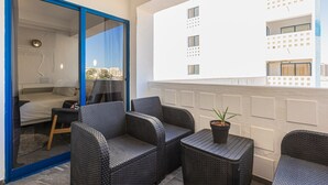 Een balkon met rieten meubilair, een tafeltje met een plant en uitzicht op een wit gebouw met meerdere ramen.