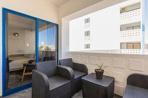 Un balcón con muebles de mimbre, una pequeña mesa con una planta y vistas a un edificio blanco con varias ventanas.