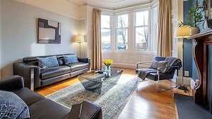 Ein modernes Wohnzimmer mit Sofa, Sessel und Couchtisch, großen Fenstern mit Vorhängen und einem Kamin.