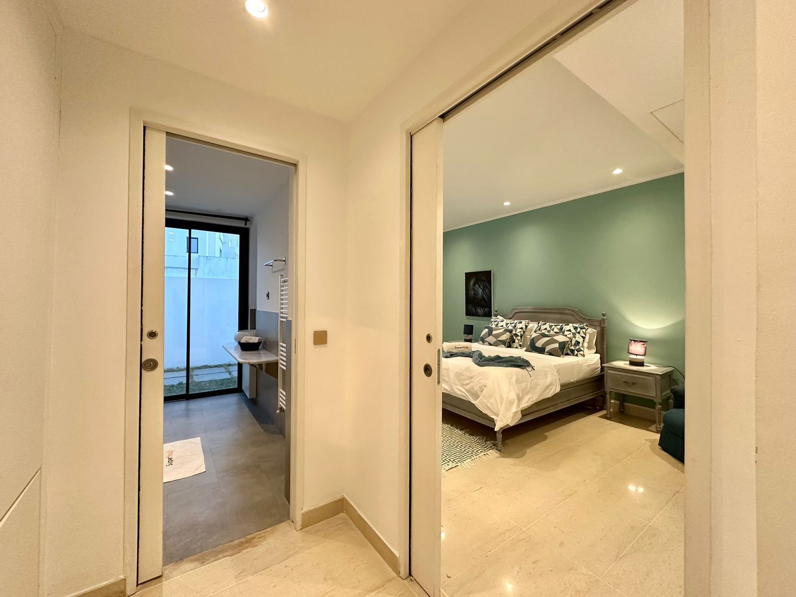 A modern bedroom with a bed, bedside tables, and a view through an open door to a balcony.