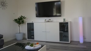 Un salon moderne doté d’un téléviseur à écran plat, d’un meuble avec des portes en verre, d’une plante en pot et d’un luminaire cylindrique.