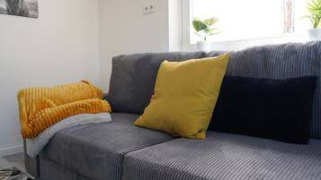 Un sofá gris con un cojín amarillo y uno negro, una manta amarilla y una planta en el alféizar de una ventana.