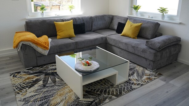 Un salon moderne avec un canapé d’angle gris, une table basse en verre et un tapis à motifs.