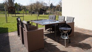 Eine Terrassengruppe im Freien mit Korbmöbeln, einem Grill und einem Spielhäuschen im Hintergrund.