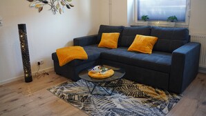 Ein Wohnzimmer mit einem dunkelgrauen Sofa, zwei gelben Kissen, einem runden Couchtisch, einem gemusterten Teppich, einem Wandbild und einem Fenster mit Jalousien.