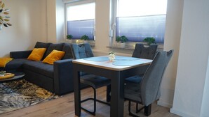 Ein modernes Wohnzimmer mit einem dunkelgrauen Sofa, passenden Sesseln und einem Holzesstisch mit Stühlen. Auf der Fensterbank stehen Blumentöpfe und es gibt eine Wanddekoration.